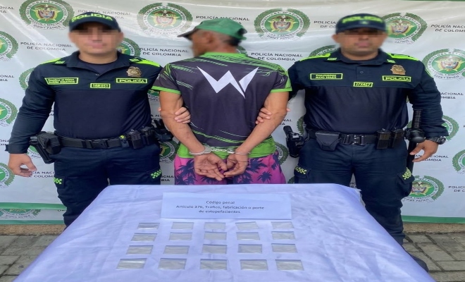Dos funcionarios policiales con un ciudadano capturado frente a pendón institucional, frente a una mesa donde esta el material incautado