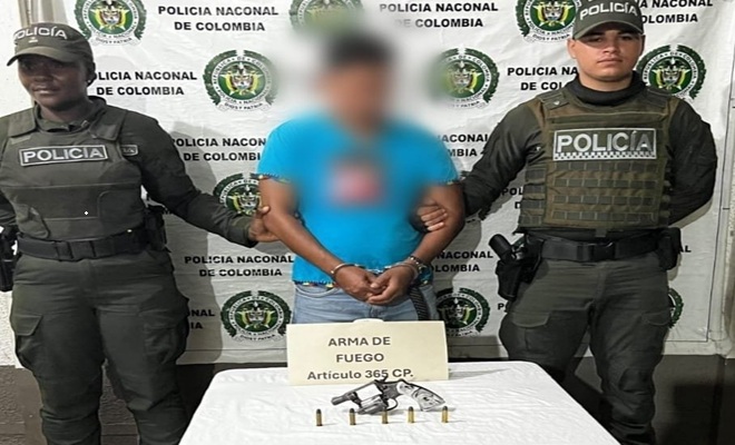 Uniformados de la Policía Nacional custodian a una persona capturada por el presunto delito de fabricación, tráfico, porte o tenencia de armas de fuego. En una mesa se observa un revólver, munición y un aviso con la leyenda “Arma de Fuego Artículo 365 C.P.”, elementos incautados durante el procedimiento realizado en el municipio de Atrato.