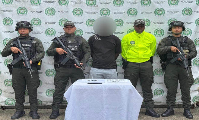 Uniformados de la Policía Nacional presentan a una persona capturada mediante orden judicial en Santa Rita de Iró, Chocó. En el procedimiento participaron integrantes del GAULA y unidades policiales. Sobre una mesa se observa un teléfono celular incautado como elemento material probatorio. Al fondo se aprecia un pendón institucional de la Policía Nacional de Colombia.