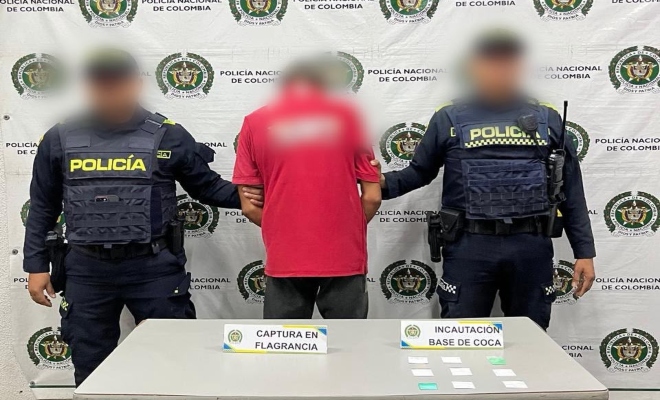 Dos funcionarios policiales con un ciudadano capturado frente a ellos una mesa con material incautado y de fondo el pendón institucional  