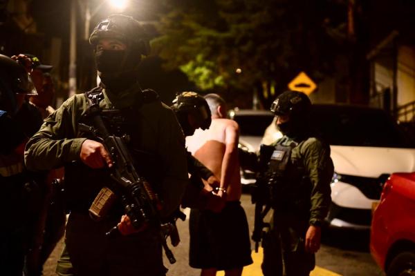 Escena nocturna en una vía urbana donde varias personas con equipo táctico se encuentran junto a vehículos estacionados; en el centro hay una persona con las manos sujetas detrás de la espalda mientras es acompañada por personal uniformado