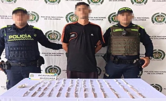 Dos funcionarios policiales con un ciudadano capturado frente a pendón institucional, frente a ellos una mesa sobre la cual están los materiales incautados