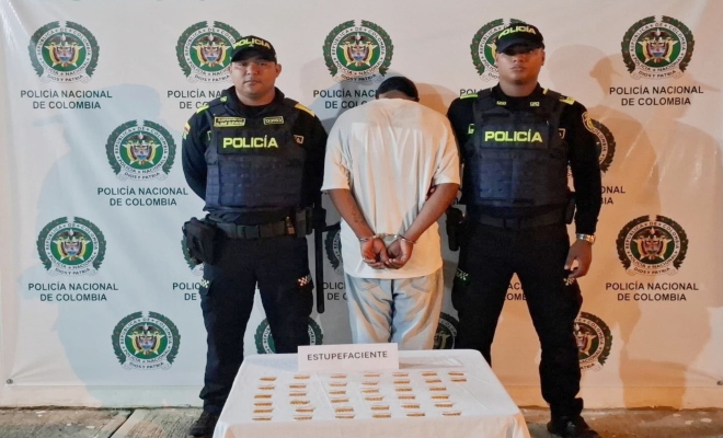 Dos funcionarios policiales con un ciudadano capturado frente a pendón institucional, frente a ellos una mesa con material incautado