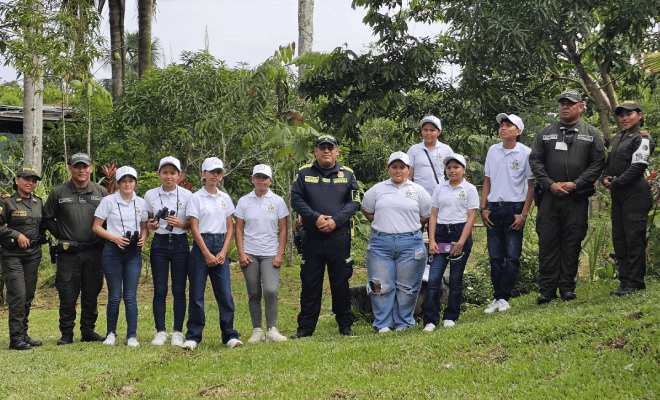 Cinco policías participan en una entrevista junto a ocho personas de civil vestidas con camisa blanca todos se encuentran de pie con árboles al fondo