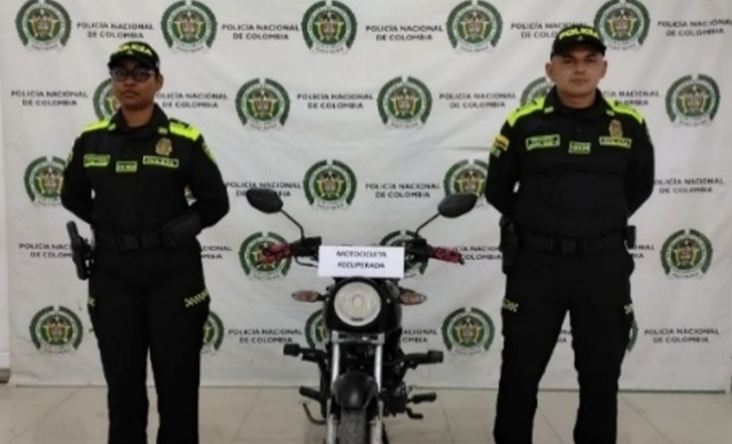 Dos funcionarios policiales junto a una motocicleta recuperada frente a un pendón institucional