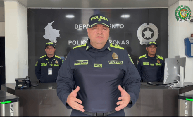 Un polic&iacute;a de pie realiza una entrevista frente a c&aacute;mara detr&aacute;s de &eacute;l se observan dos polic&iacute;as de pie a los lados hay un computador y al fondo el escudo de la Polic&iacute;a Nacional y el mapa de Colombia