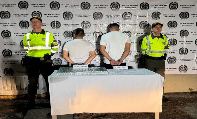 Dos personas de espaldas capturadas por el delito de tráfico de sustancias para el procesamiento de narcóticos, elementos incautados sobre una mesa con mantel blanco y dos Policías de la Seccional de Tránsito y Transporte brindando seguridad.