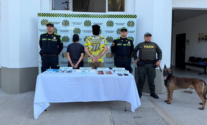 Dos personas de espalda capturadas por el delito de tráfico de estupefacientes, elementos incautados sobre una mesa con mantel blanco, tres funcionarios de la Policía brindando seguridad y un canino del grupo de estupefacientes