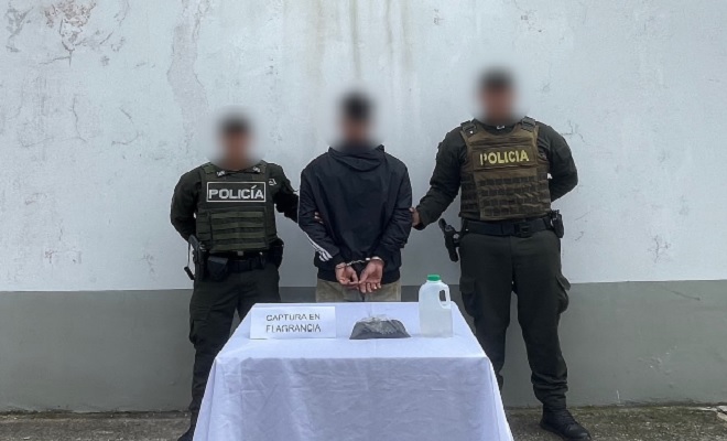 Una persona capturada acompañada por dos policías, en frente de ellos una mesa con elementos incautados 