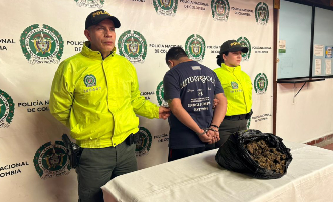 Hombre capturado en medio de dos Policías, en frente se encuentra ubicada una mesa con gramos de marihuana incautada