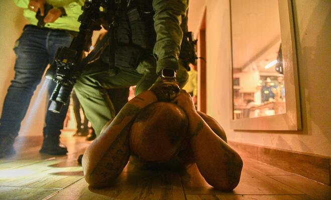  Interior con iluminación cálida donde una persona se encuentra acostada boca abajo sobre un piso de madera con las manos sujetas detrás de la cabeza, mientras una persona uniformada se sitúa encima sosteniendo un arma larga; en el fondo se observan otras personas y un espacio tipo pasillo.