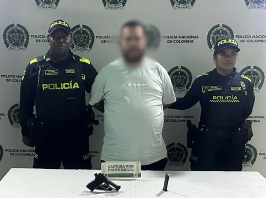 fondo institucional de la Policía Nacional de Colombia; tres personas de pie, una en el centro con las manos sujetas detrás, acompañada por dos personas uniformadas a los lados; en primer plano hay una mesa con un arma corta y un objeto cilíndrico, junto a un rótulo que dice “captura por porte ilegal"