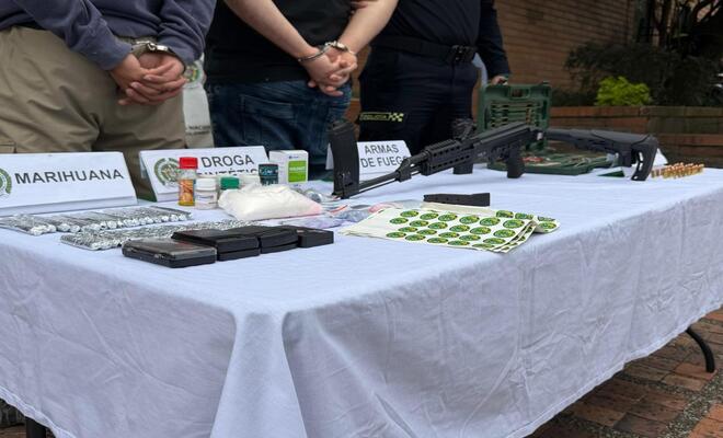 Mesa cubierta con mantel blanco donde se exhiben objetos etiquetados, incluyendo paquetes, frascos, municiones y armas largas; al fondo se observan personas con las manos sujetas detrás del cuerpo.
