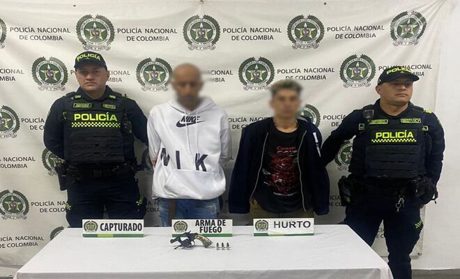 En un espacio institucional, dos personas con los rostros difuminados se encuentran de pie entre dos integrantes de la Polic&iacute;a Nacional. Frente a ellos hay una mesa con un arma de fuego, munici&oacute;n y r&oacute;tulos que indican "capturado", "arma de fuego" y "hurto". 