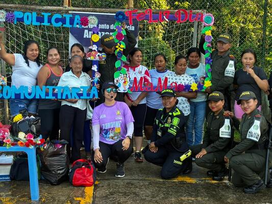 Un grupo mixto conformado por miembros de la fuerza policial y vecinos de la comunidad posan juntos de forma alegre y participativa, en un espacio al aire libre decorado con letreros y elementos coloridos que identifican la actividad. Se aprecian artículos y materiales en el suelo, lo que evidencia que se trata de una jornada de integración, convivencia y acercamiento entre la institución y la población