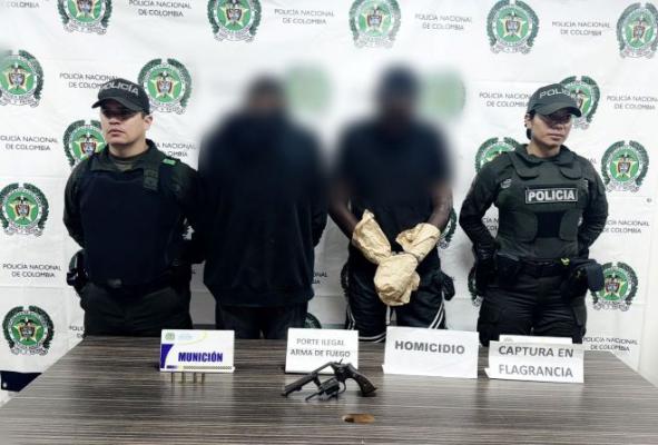 Dos polic&iacute;as al lado de dos hombres capturados por homicidio y un arma de fuego encima de una mesa
