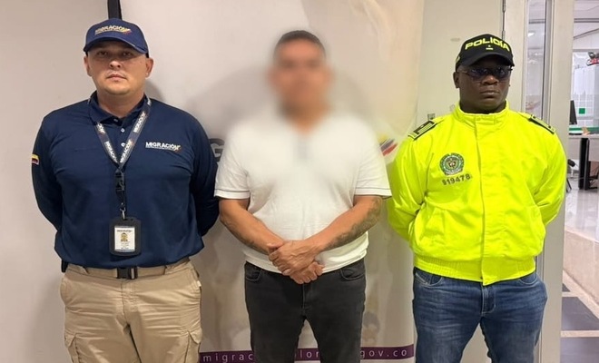 Tres personas de pie en un espacio interior frente a un panel informativo; a los lados se observan una persona con uniforme de Migración Colombia y otra con uniforme de la Policía Nacional, mientras la persona ubicada en el centro viste camiseta clara y pantalón oscuro.