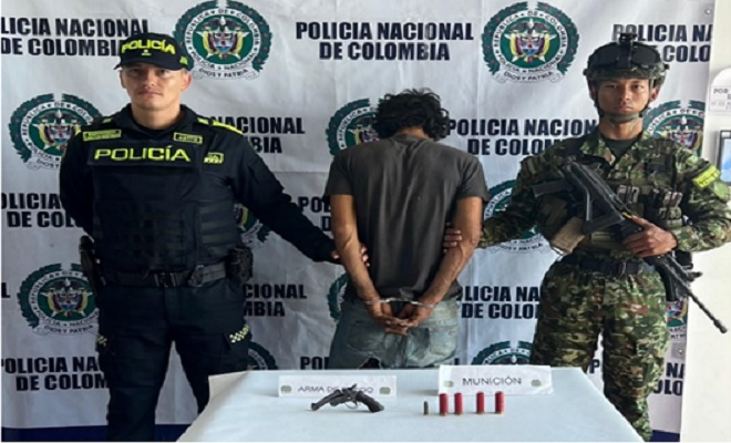 arma de fuego y proveedores incautados sobre una mesa con hombre detenido y un soldado y un policía a cada lado