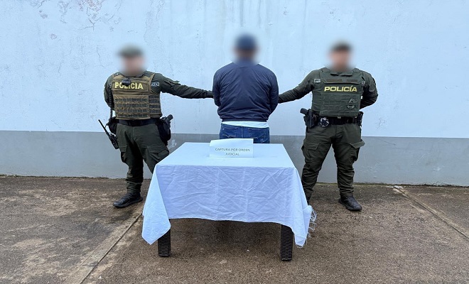 Dos policías presentan a persona capturada, en frente una mesa con mantel blanco 