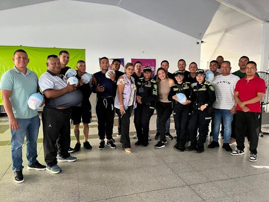 Una amplia agrupación de personas, compuesta por civiles y agentes de la policía, se reúnen al finalizar la actividad, muestran expresiones de satisfacción y alegría, y varios de ellos sostienen objetos redondos en sus manos, bajo la organización y el nombre identificativo del programa