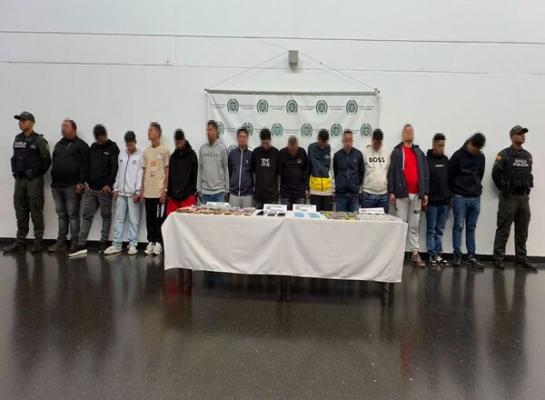 Espacio interior con fondo institucional de la Policía Nacional de Colombia donde una fila de varias personas está de pie frente a una mesa cubierta con un mantel blanco; sobre la mesa hay objetos organizados en filas y dos personas uniformadas se ubican en los extremos.