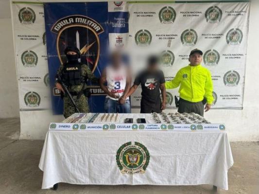 un policía y GAULA militar al lado de dos hombres capturados con municiones estupefacientes celulares